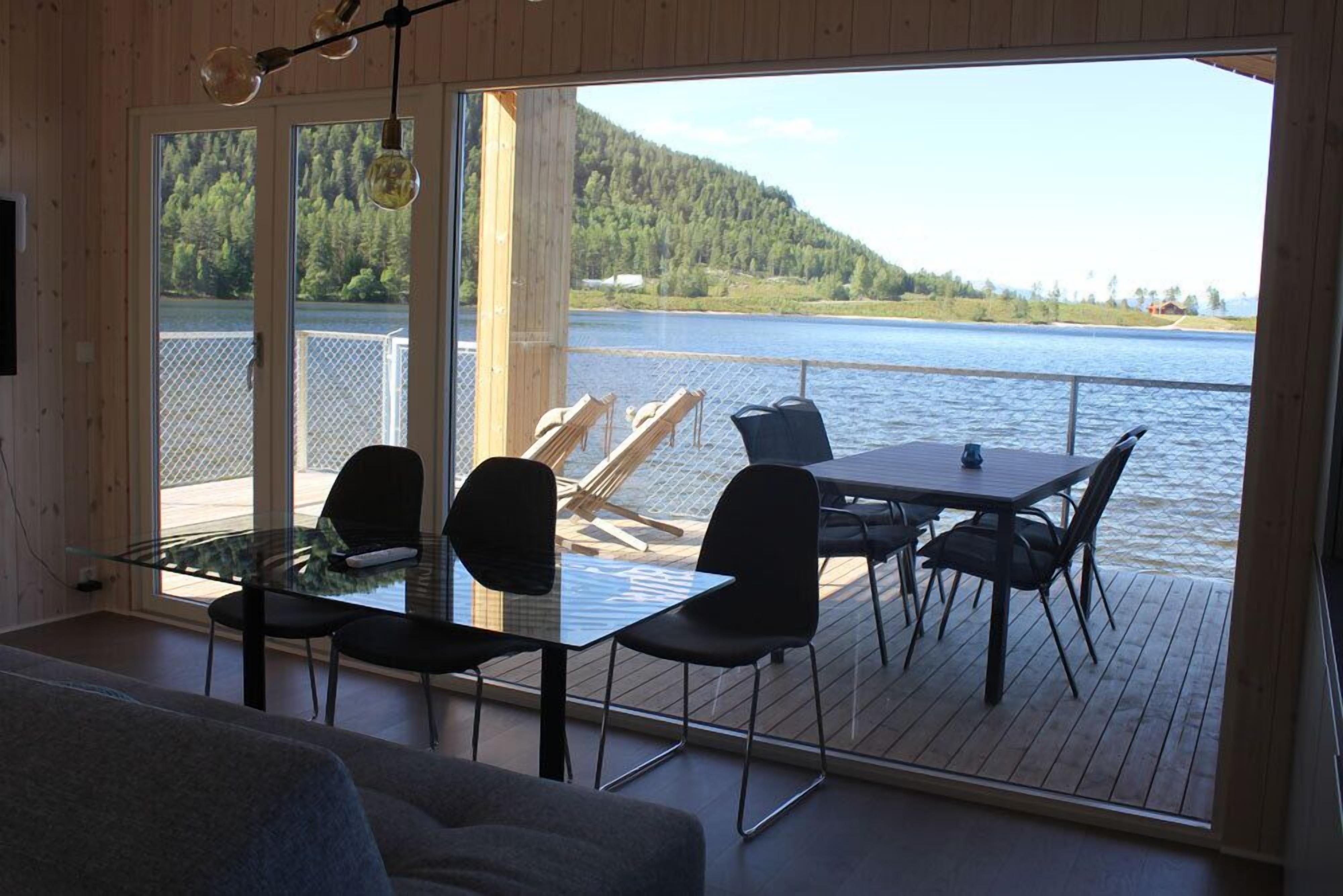 Una habitación con una mesa y sillas de vidrio, una terraza de madera y vistas a un lago y un bosque.