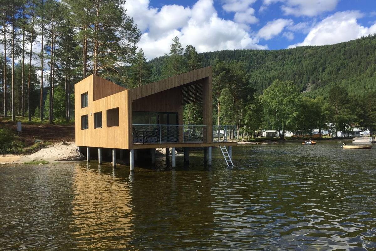 A wooden cabin on stilts over a lake, with a deck and railing, surrounded by forest and boats.