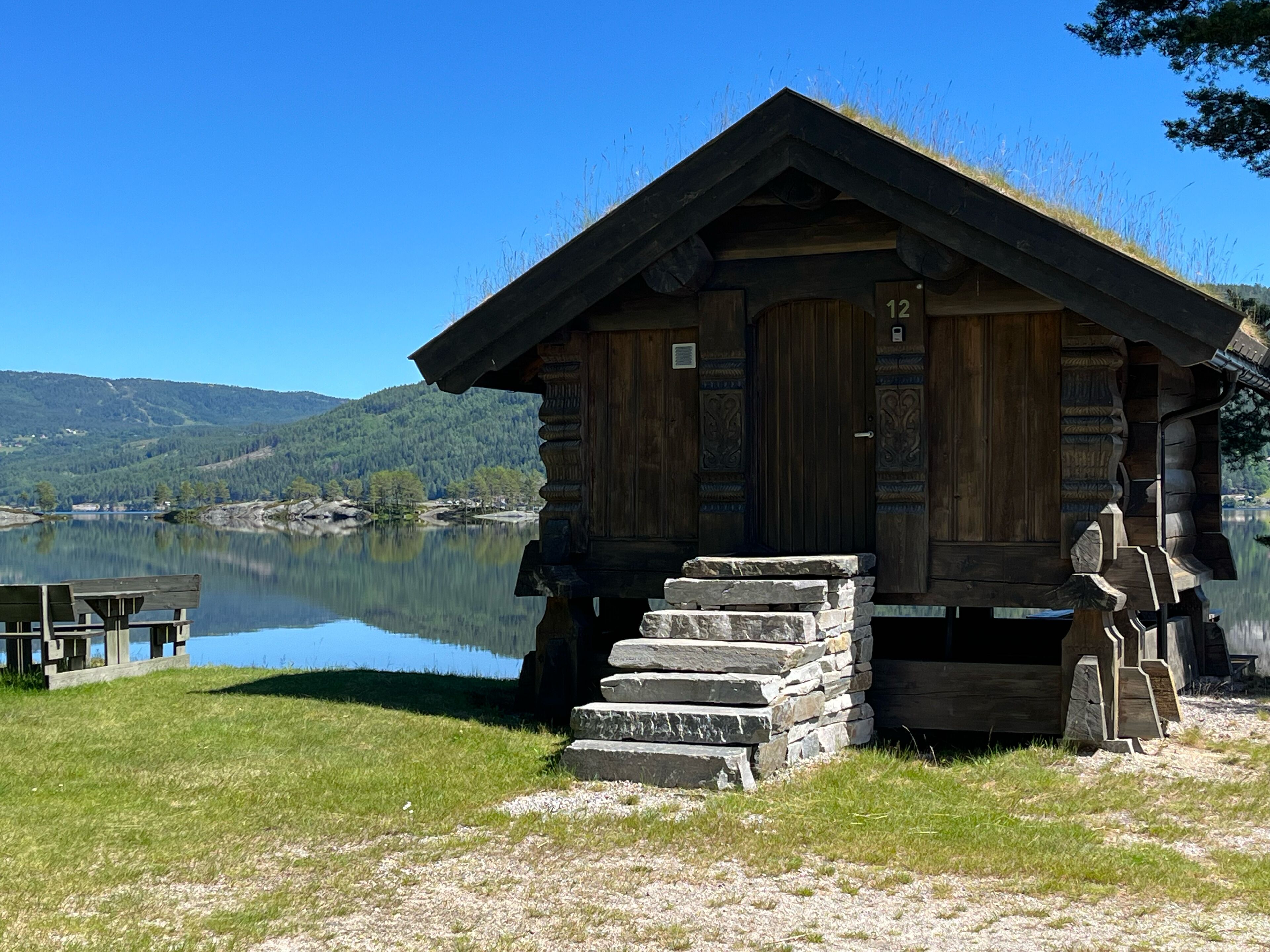 Una cabaña de madera con una escalera de piedra que sube a una puerta, ubicada junto a un lago tranquilo con un paisaje boscoso de fondo.
