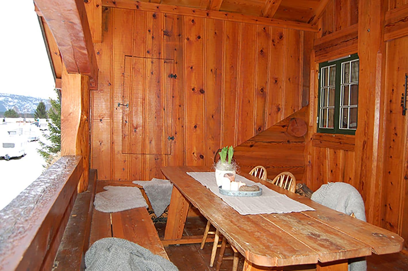 Une cabane en bois avec une table et des bancs en bois, une fenêtre avec des volets verts, et une vue sur un parking et des montagnes.