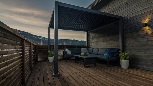 Terraza de madera con zona de estar cubierta, una planta en maceta y vistas a montañas nevadas.