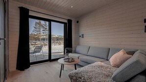 A modern living room with a grey sofa, a fluffy rug, and a small round table. A large window offers a view of a snowy landscape.