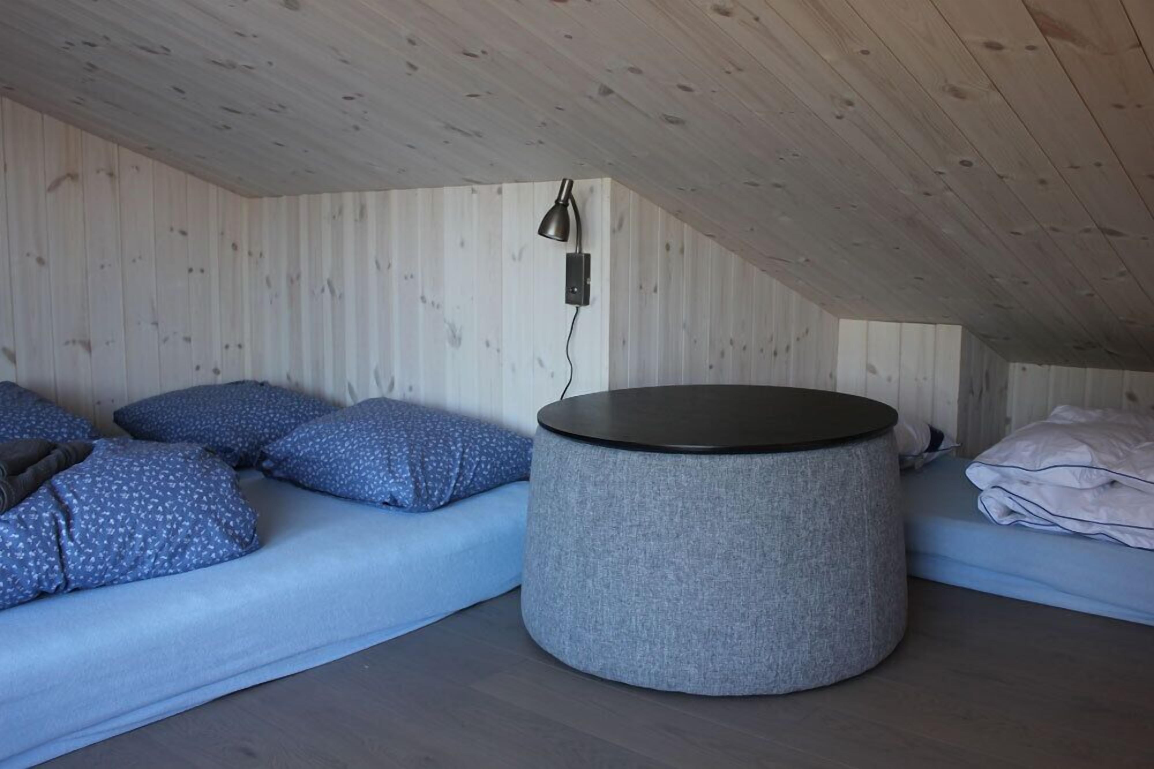 Una habitación con techo de madera, un otoman gris redondo, un sofá azul con cojines estampados y una cama blanca con una manta doblada.