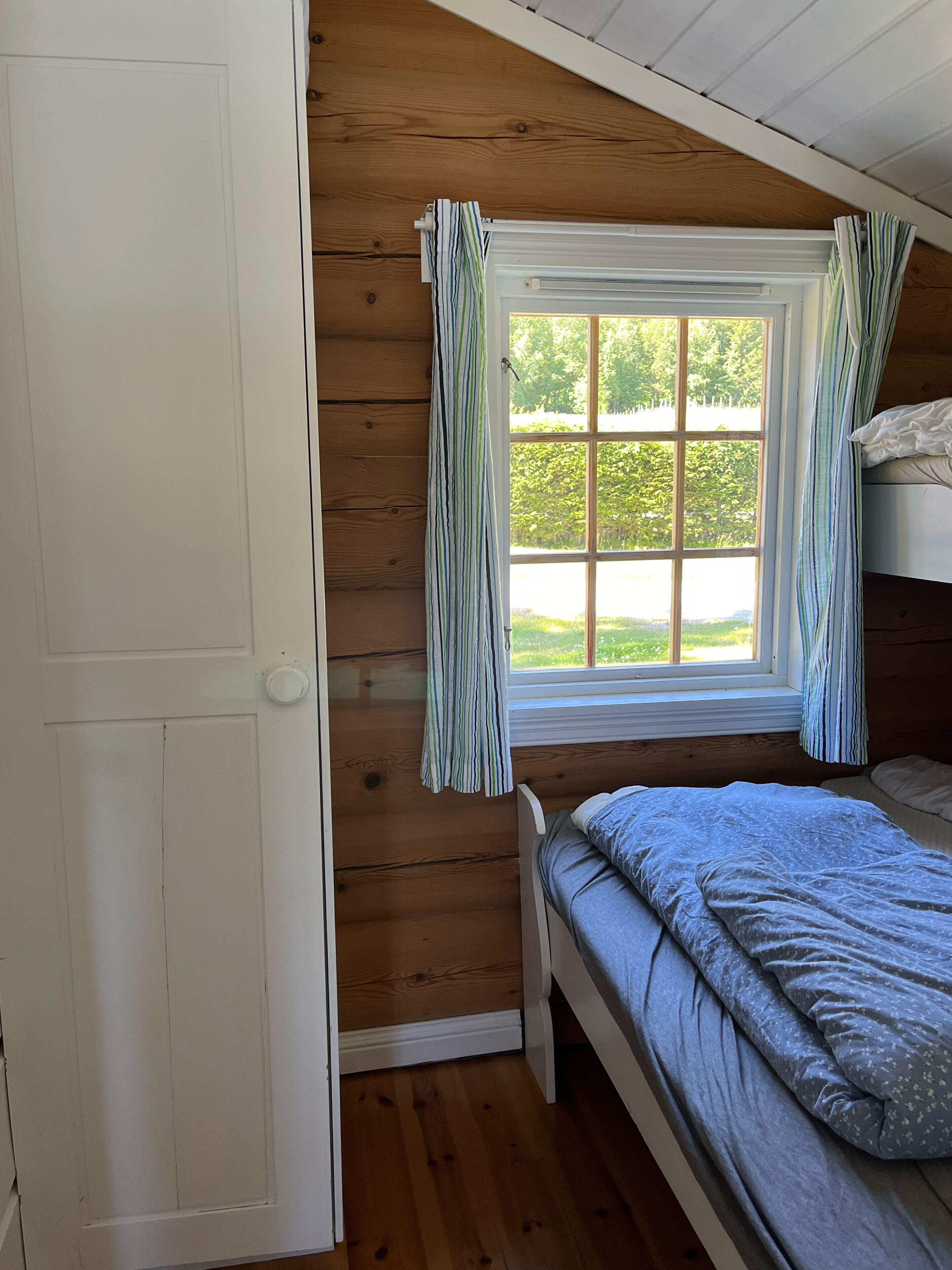 Una habitación tipo cabaña con interior de madera, una ventana con cortinas, una cama y una puerta.
