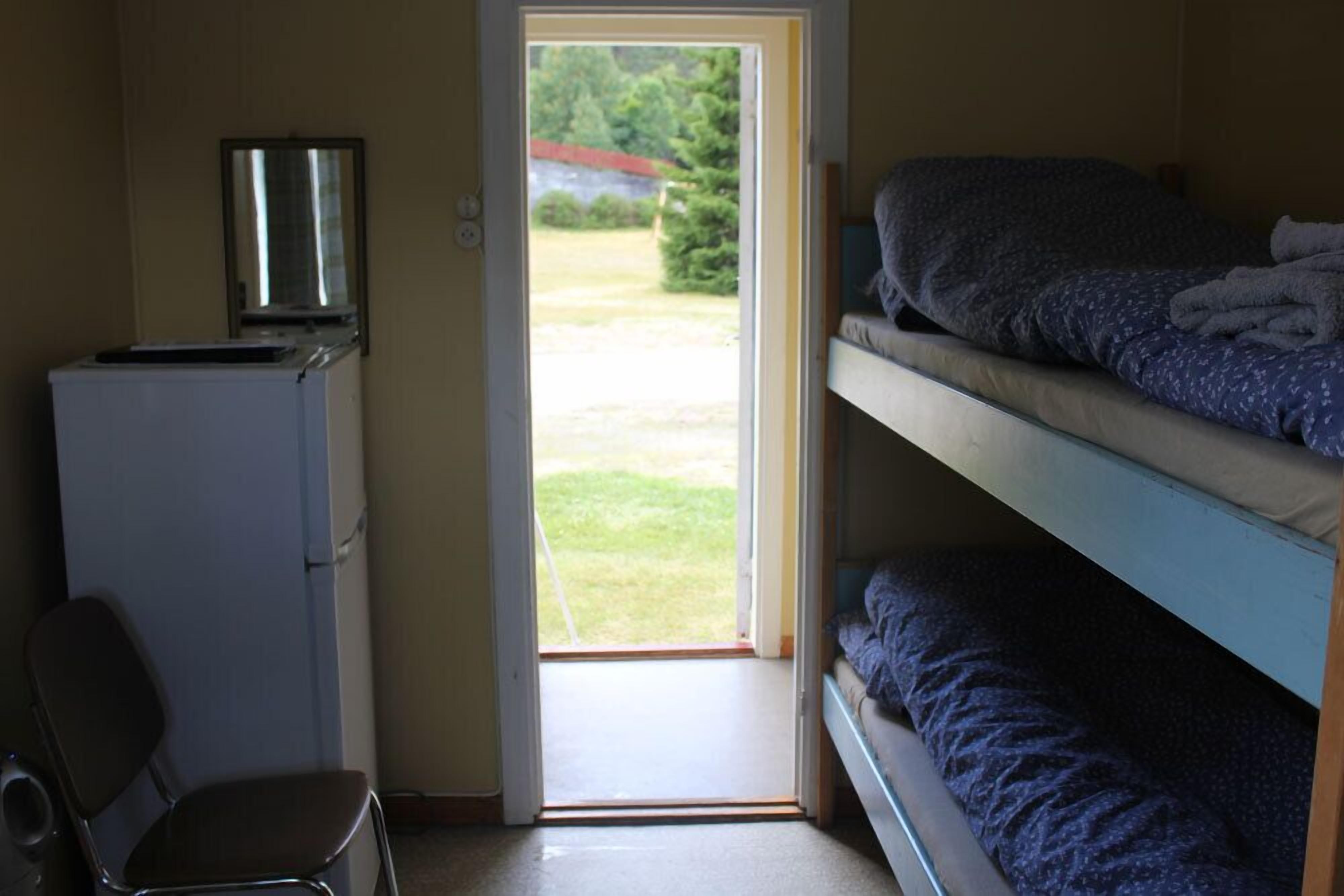 A dormitory room with bunk beds, a desk with a computer, and a door leading to a grassy area.