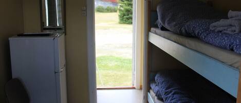 A dormitory room with bunk beds, a desk with a computer, and a door leading to a grassy area.