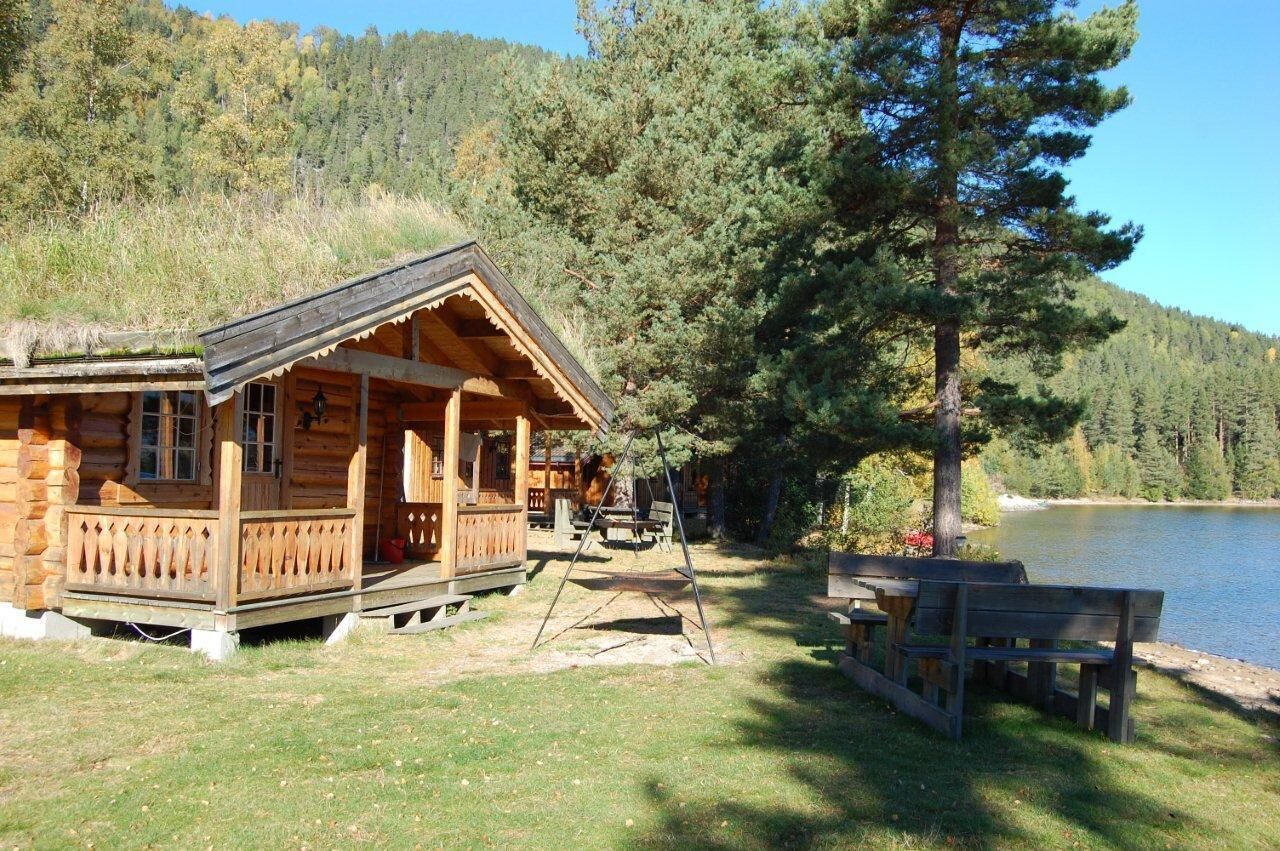 A wooden cabin with a porch and a swing set, located near a lake surrounded by trees.