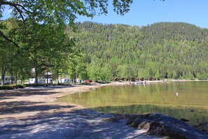 Caserío junto al lago con tiendas de campaña y cabañas, rodeado de un bosque denso y un cielo despejado.