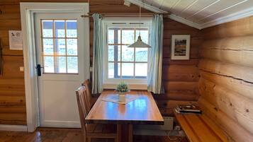Et interiør af en træhytte med et spisebord, stole, en bænk, et vindue med gardiner og en loftlampe.