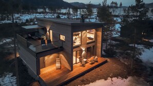 A modern, two-story cabin with a glass balcony, wooden deck, and a view of a snowy landscape.