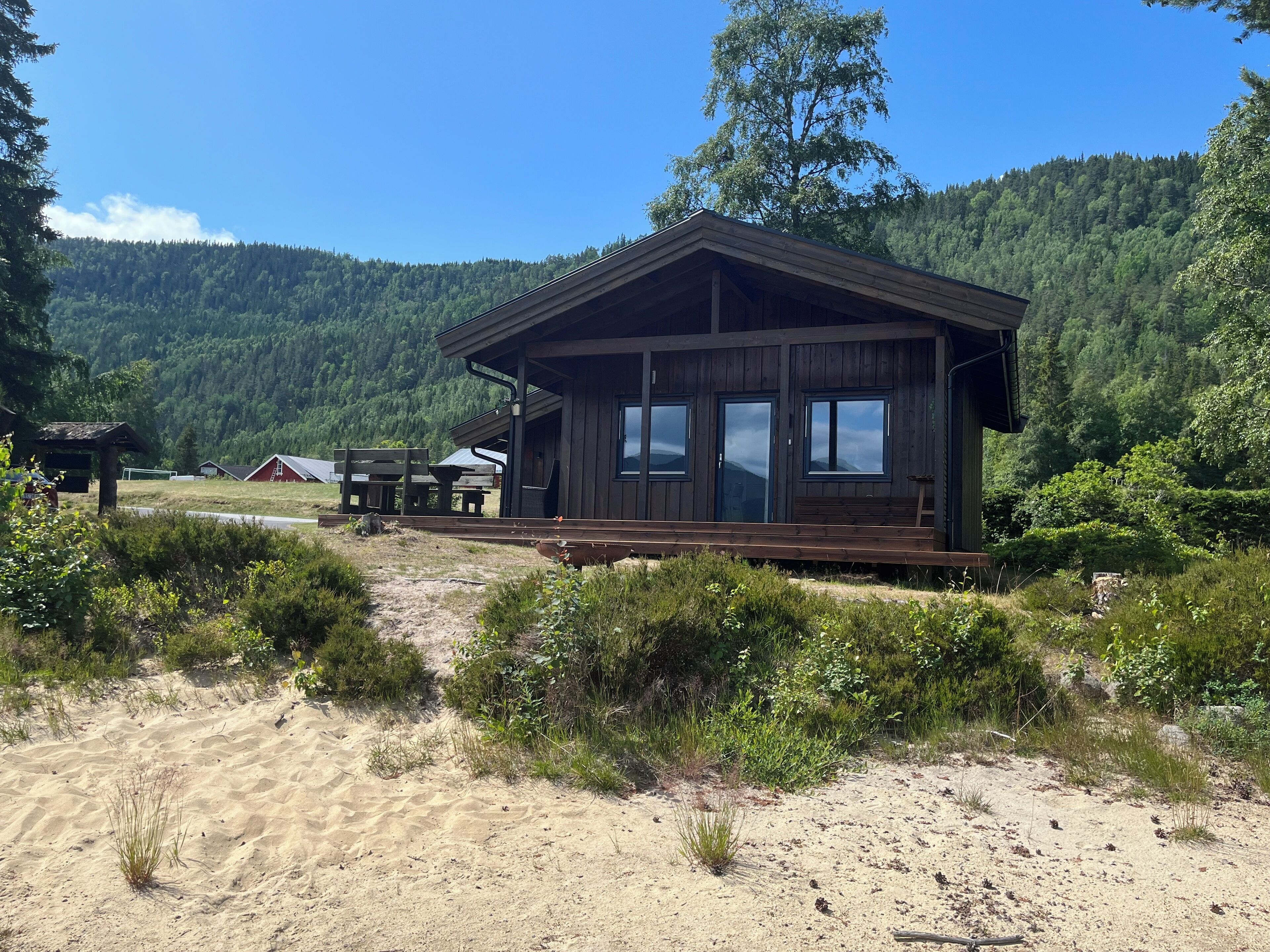 A wooden cabin with a porch, surrounded by a forested area and a sandy path.