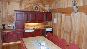 A rustic cabin interior with a wooden dining table, red cabinets, a kitchen area, and a mounted deer head.