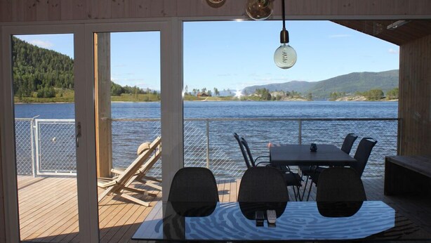 A wooden deck with a table and chairs overlooking a lake, with a view of hills and a fence in the background.