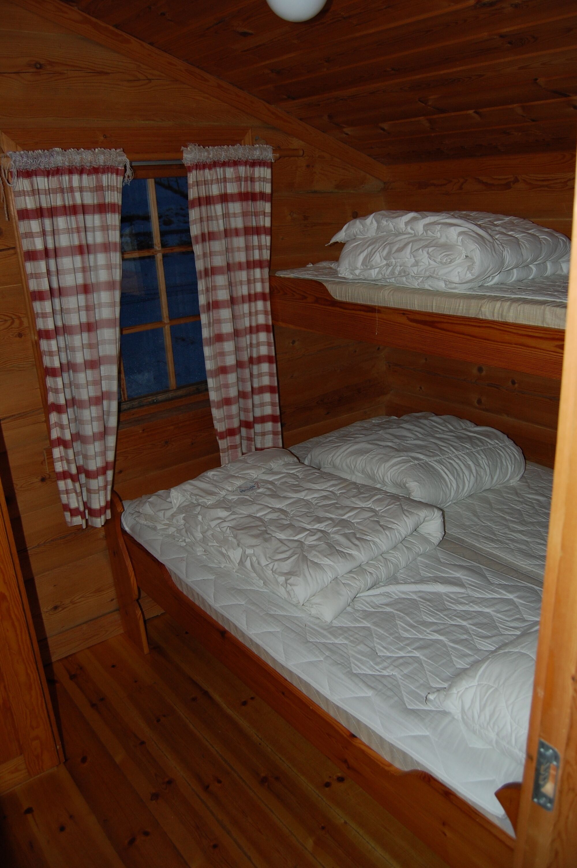A cabin with a wooden interior, a bunk bed with white bedding, and plaid curtains on a window.
