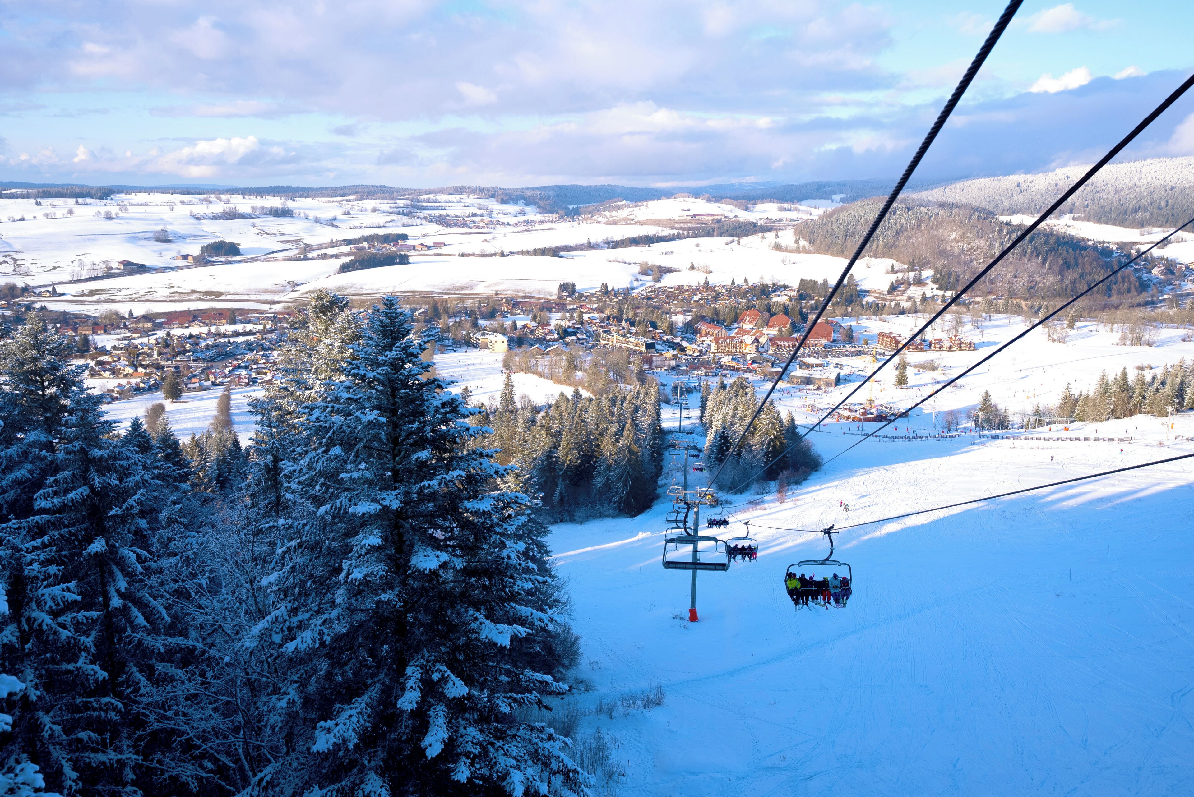 A ski lift with cabins moving over a snowy landscape, surrounded by trees and a small town below.