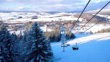 A ski lift with cabins moving over a snowy landscape, surrounded by trees and a small town below.