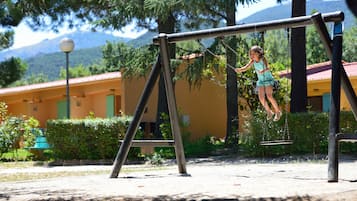 Children can play on the playground with a swing set surrounded by lush greenery and trees.