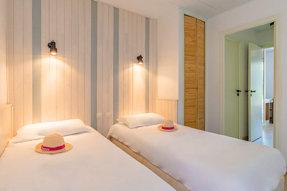 Two single beds with white linens and a hat on each, wooden paneling on the walls, and a wooden door in the background.