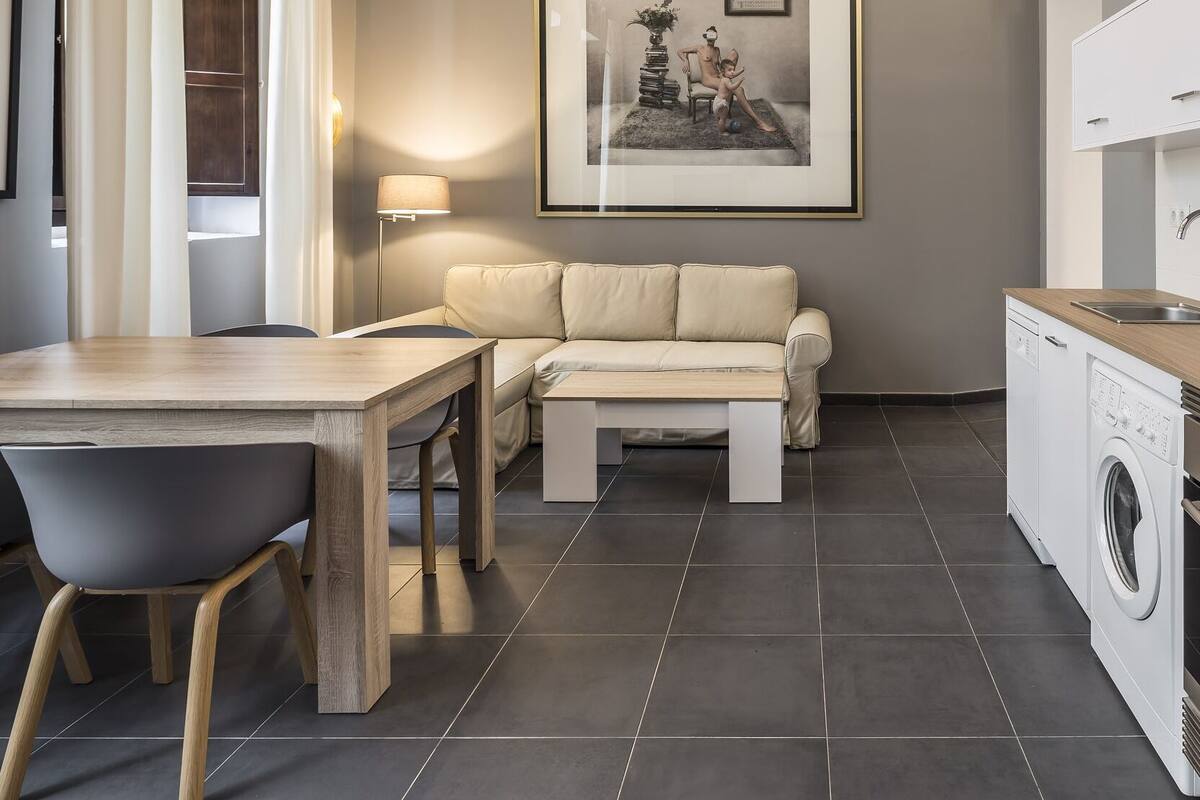 Una cocina moderna con lavadora integrada, un comedor con mesa y sillas, y una sala de estar con sofá y un cuadro en la pared.