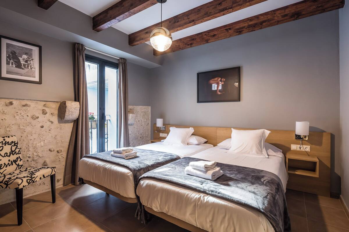 Habitación de hotel con dos camas, techo de madera, un ventanal grande con cortinas, un cuadro colgado en la pared y una mesita de noche con lámpara.