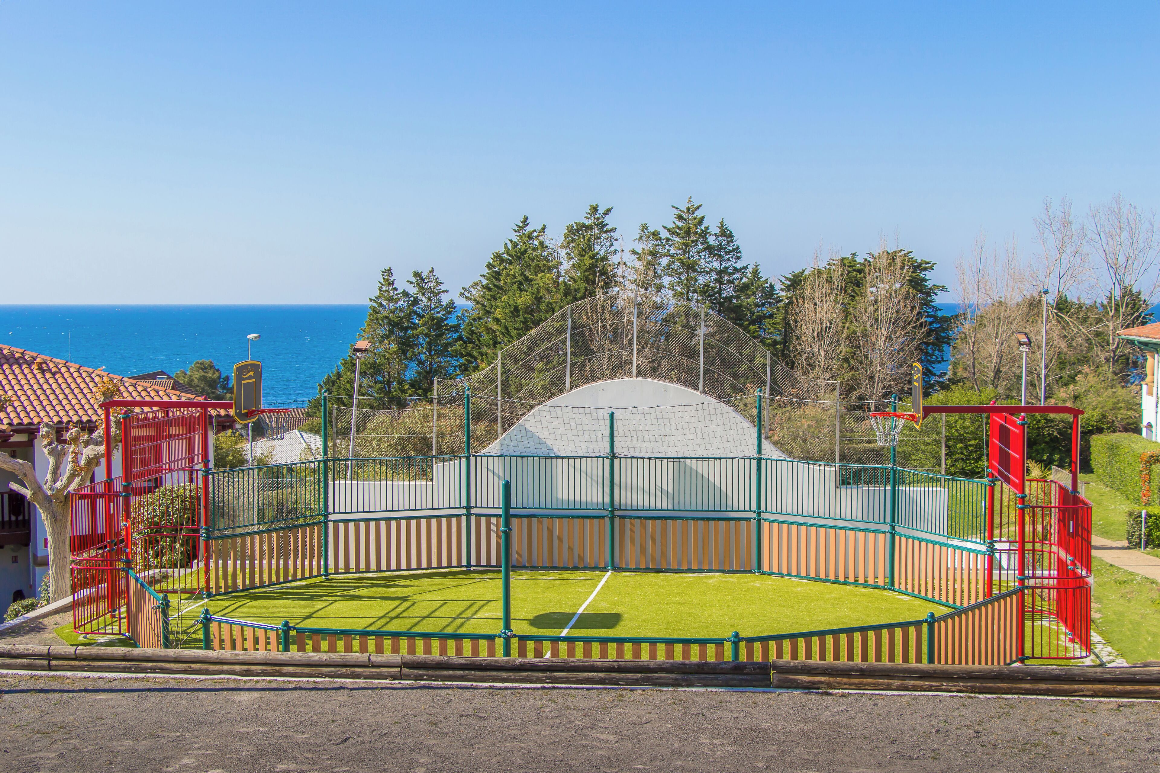 Una cancha deportiva con una red, rodeada por una valla, y vista al mar en el fondo.