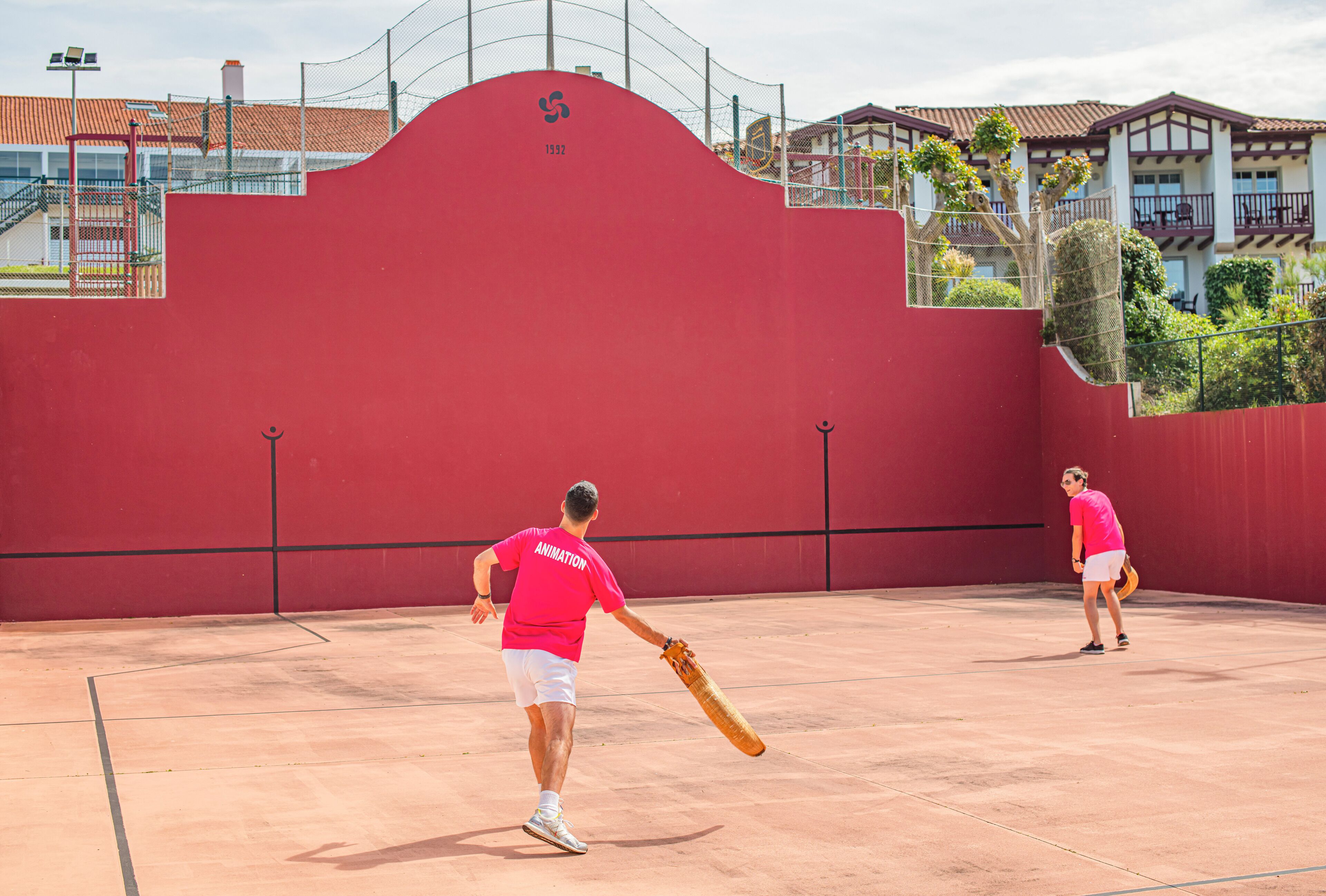 A tennis court with a red wall and a building with balconies in the background.