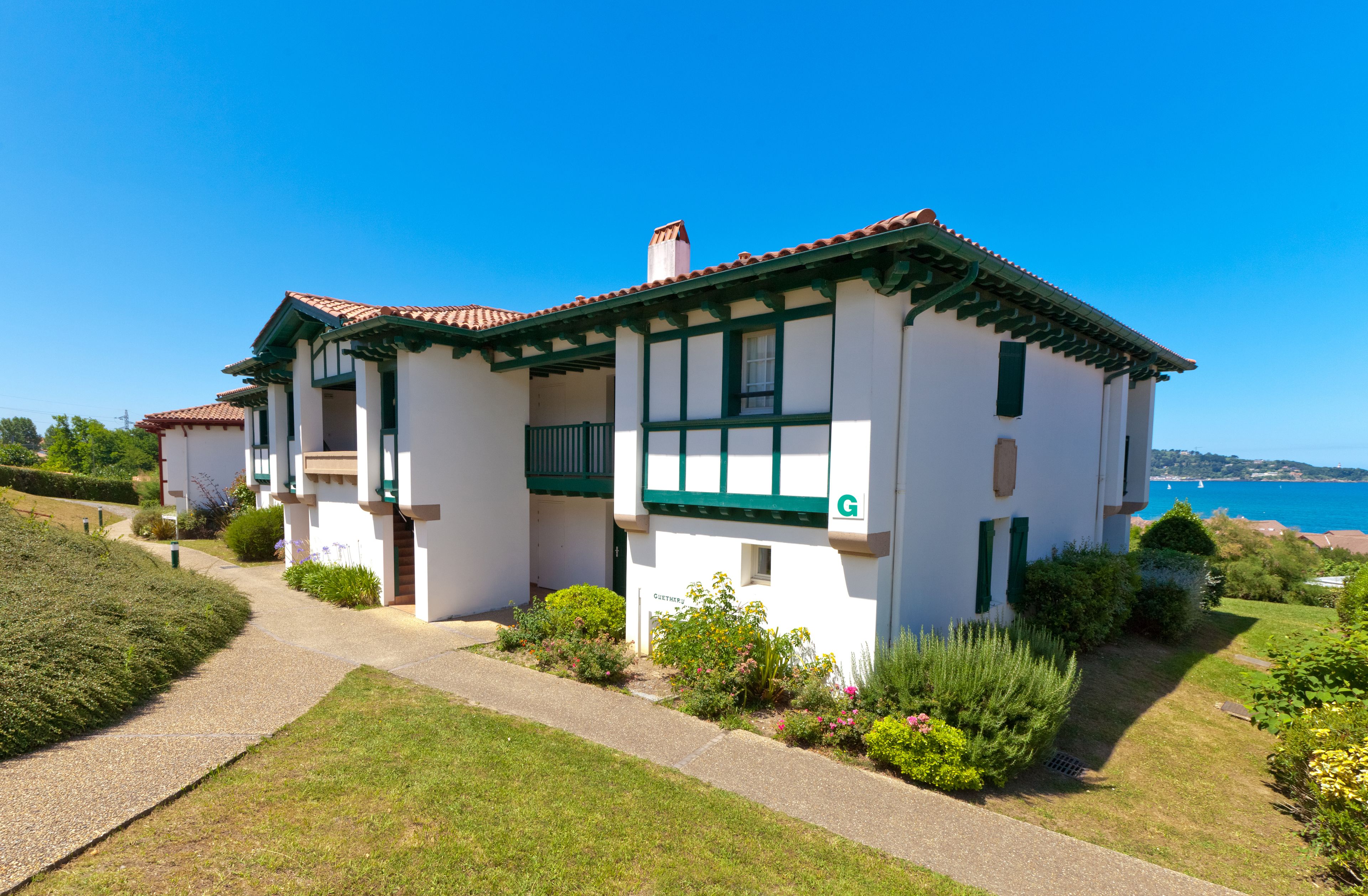 A two-story house with a white exterior and green trim, a visible 'G' logo, and a well-maintained garden with a pathway leading to the entrance.