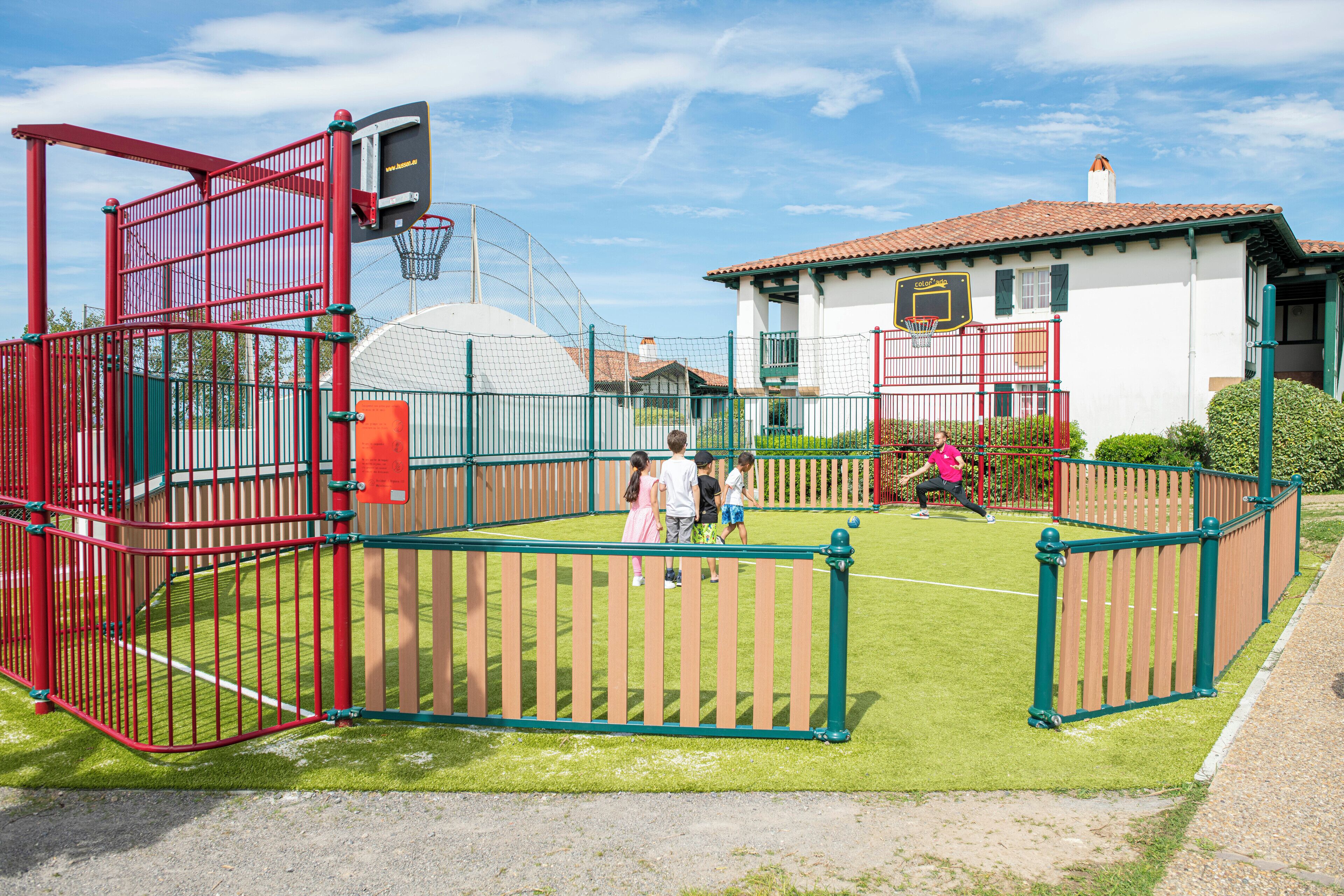 Una cancha deportiva cercada con un aro de baloncesto, un pequeño edificio con techo rojo y un cielo despejado.