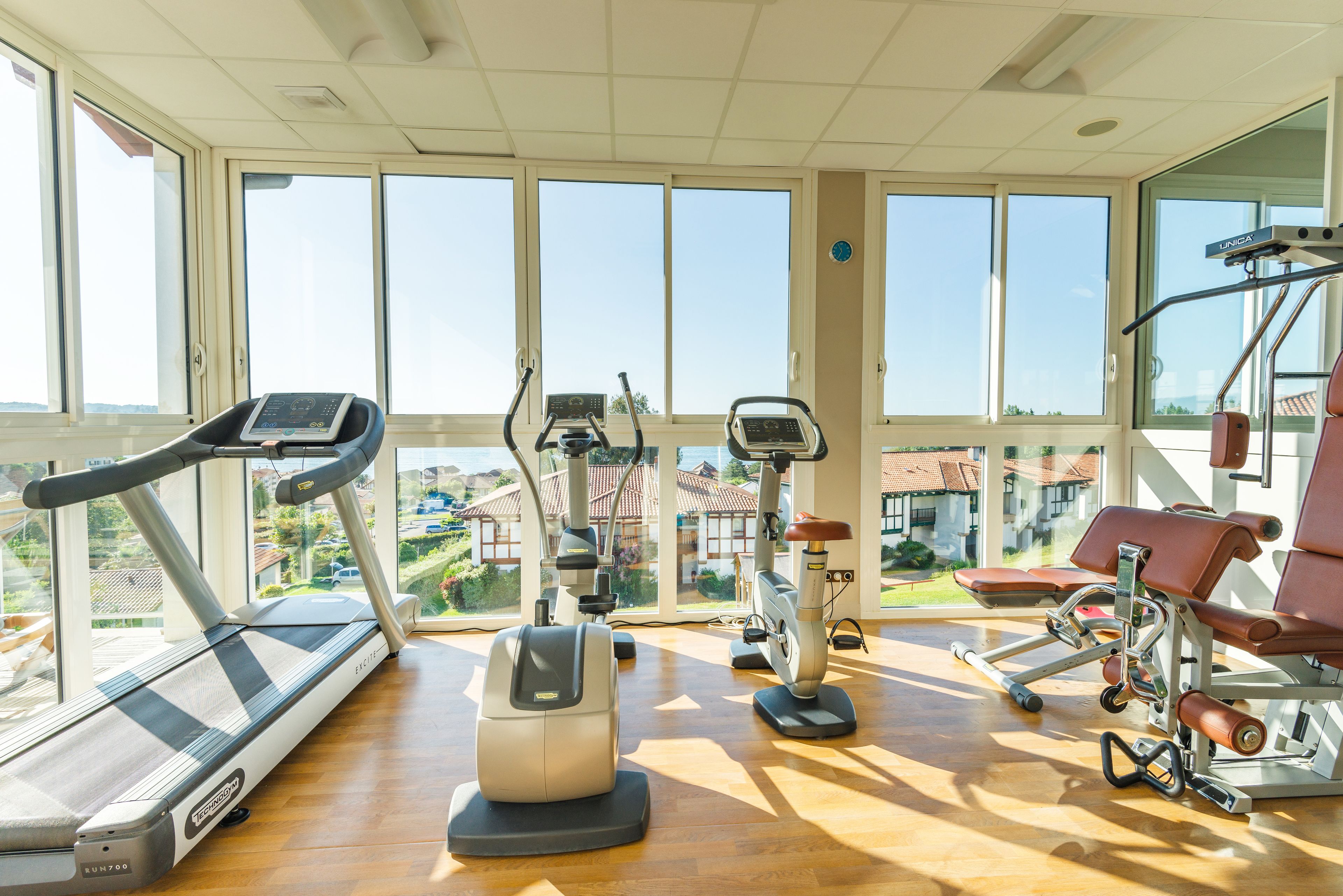 Un gimnasio con diverso equipo de ejercicio, ventanas amplias y vistas al exterior.