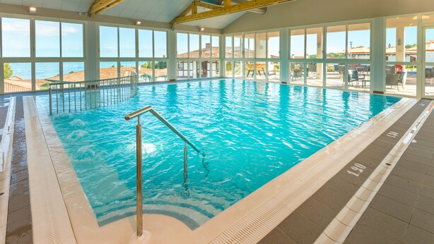 Ein Innenpool mit klarem, blauem Wasser, Holzbalken und großen Fenstern mit Blick auf das Meer und Gebäude.