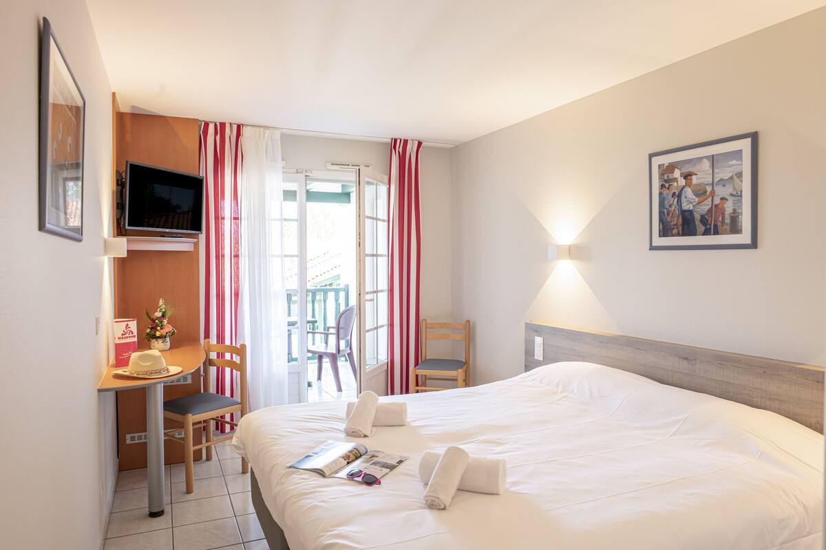 Habitación de hotel con cama, escritorio, silla, televisor y ventana con cortinas.