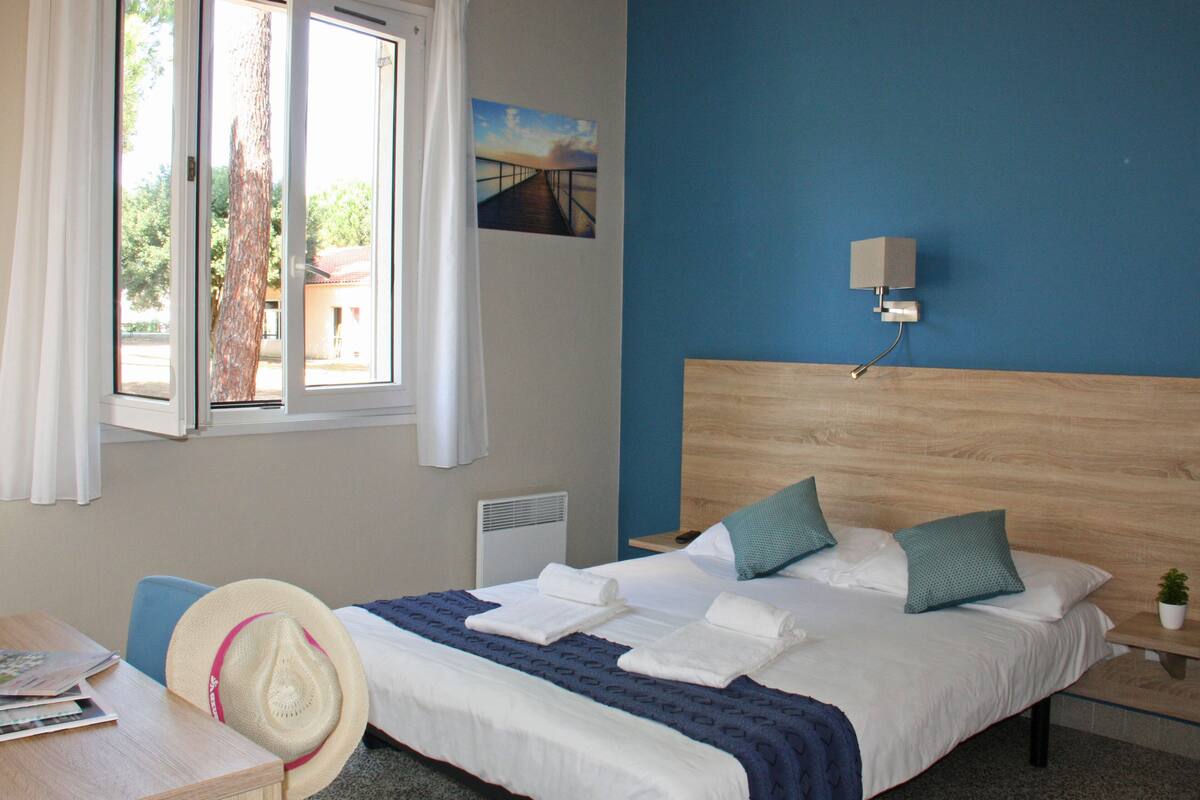Une chambre d’hôtel comprenant un lit, un bureau, une chaise, une fenêtre avec des rideaux, une lampe fixée au mur et un tableau accroché au mur.