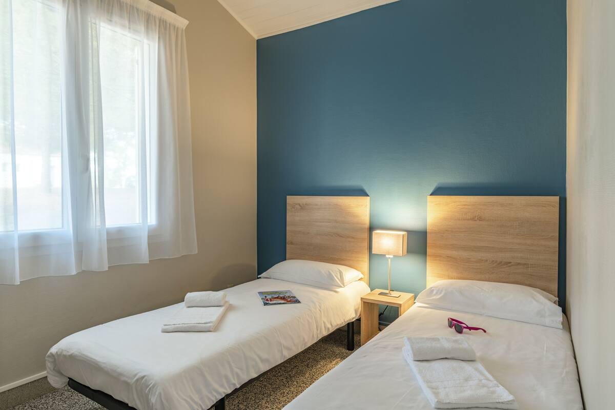 Une chambre d’hôtel avec deux lits, une table de chevet avec une lampe et une fenêtre avec des rideaux légers.