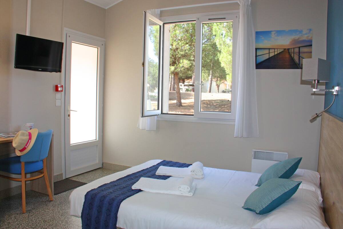 Une chambre d’hôtel avec un lit, un bureau, une chaise, une télévision et une fenêtre donnant sur des arbres et un coucher de soleil.