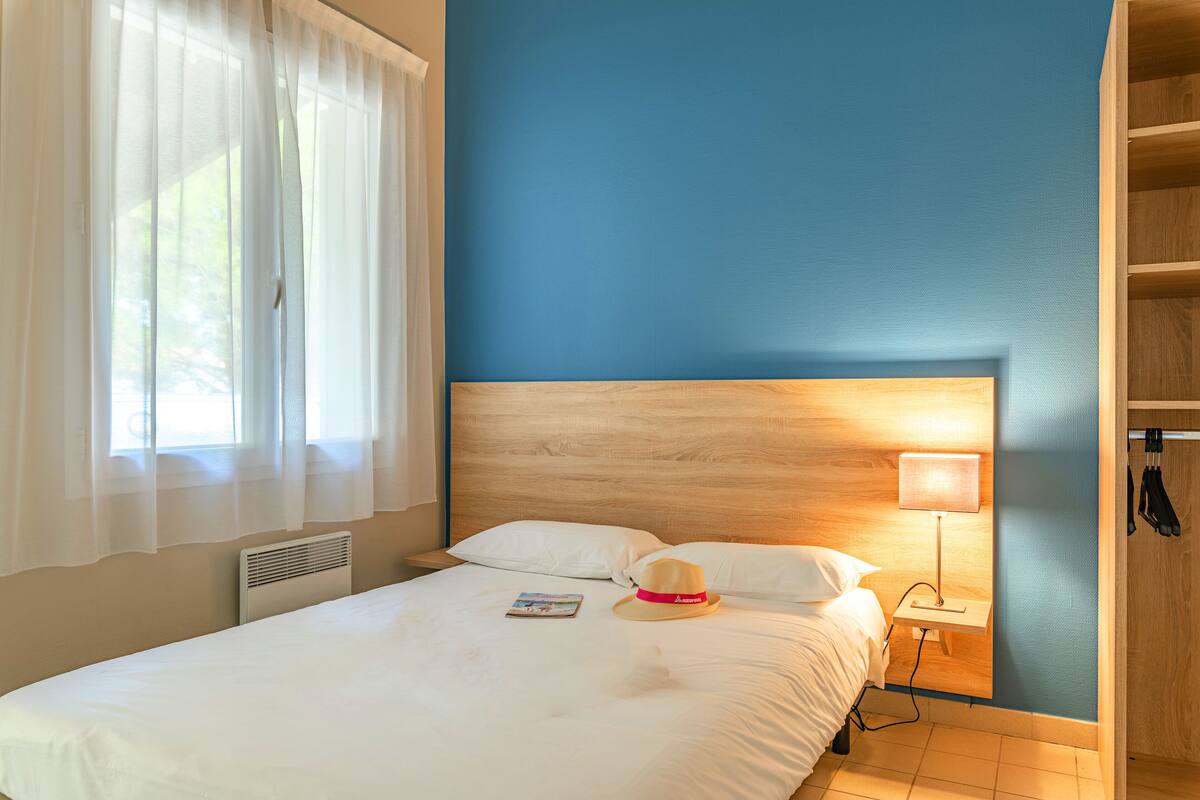 Une chambre d’hôtel avec une tête de lit en bois, une lampe de chevet, un couvre-lit blanc et une fenêtre avec des rideaux légers.