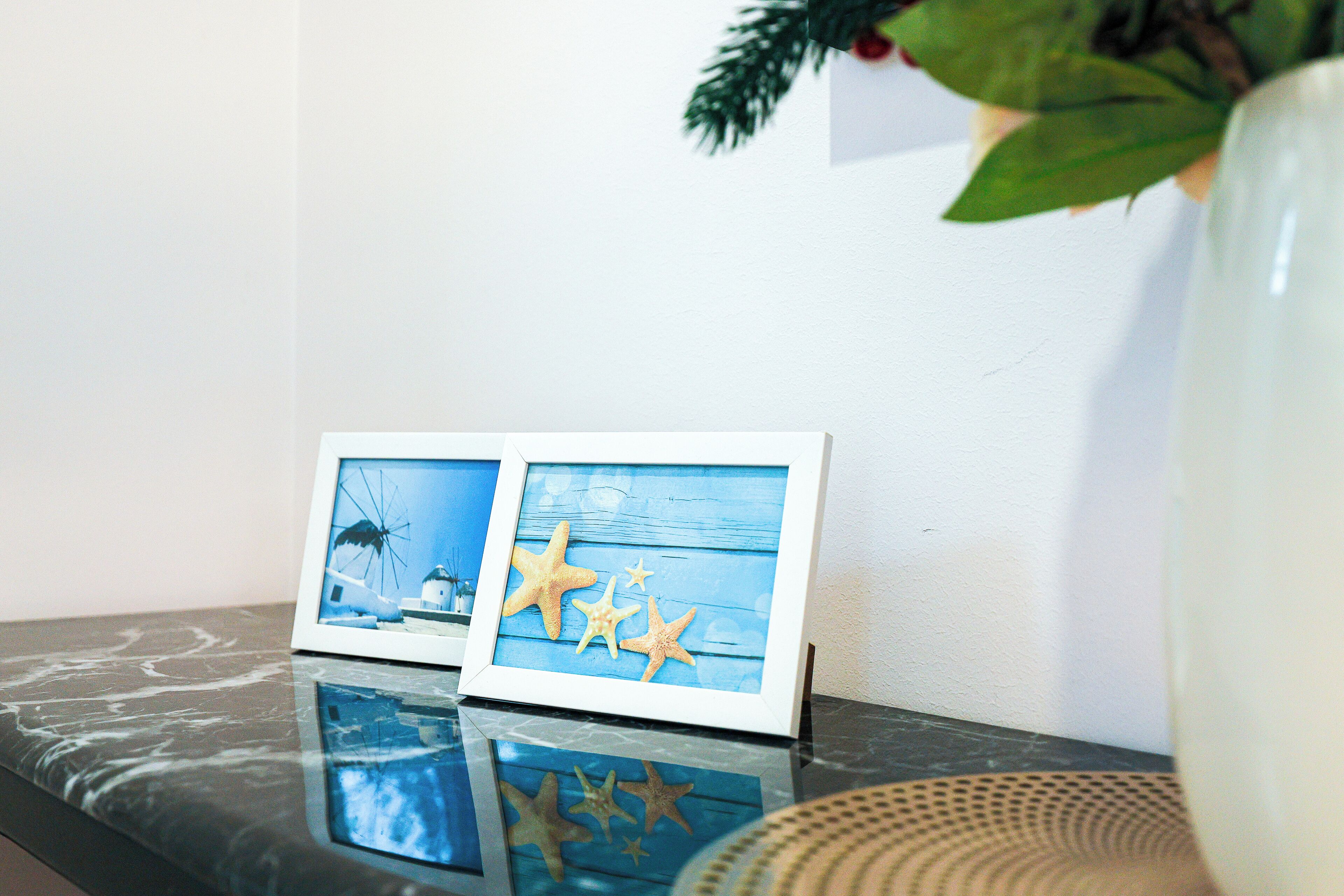 Une photographie encadrée représentant un paysage côtier avec un bateau et un moulin à vent, ainsi qu'un vase contenant une plante, le tout posé sur une surface réfléchissante.