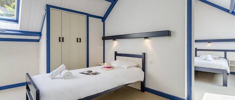 A single bed with a white mattress and pillows, a small bedside table with a book and a hat, and a window with blinds.