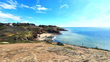 A coastal landscape with a sandy beach, rocky terrain, and a few houses on a hillside.