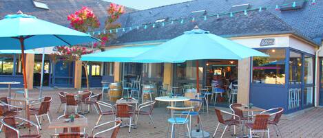 An outdoor patio with tables and chairs, umbrellas, and a building with a sign that reads "Bar".