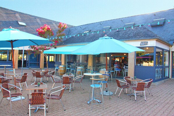 Un patio al aire libre con mesas y sillas, sombrillas y un edificio con un letrero que dice "Bar".
