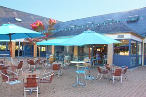 An outdoor patio with tables and chairs, umbrellas, and a building with a sign that reads "Bar".