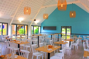 A spacious dining area with white chairs and tables, orange hanging lights, and large windows offering a view of greenery outside.
