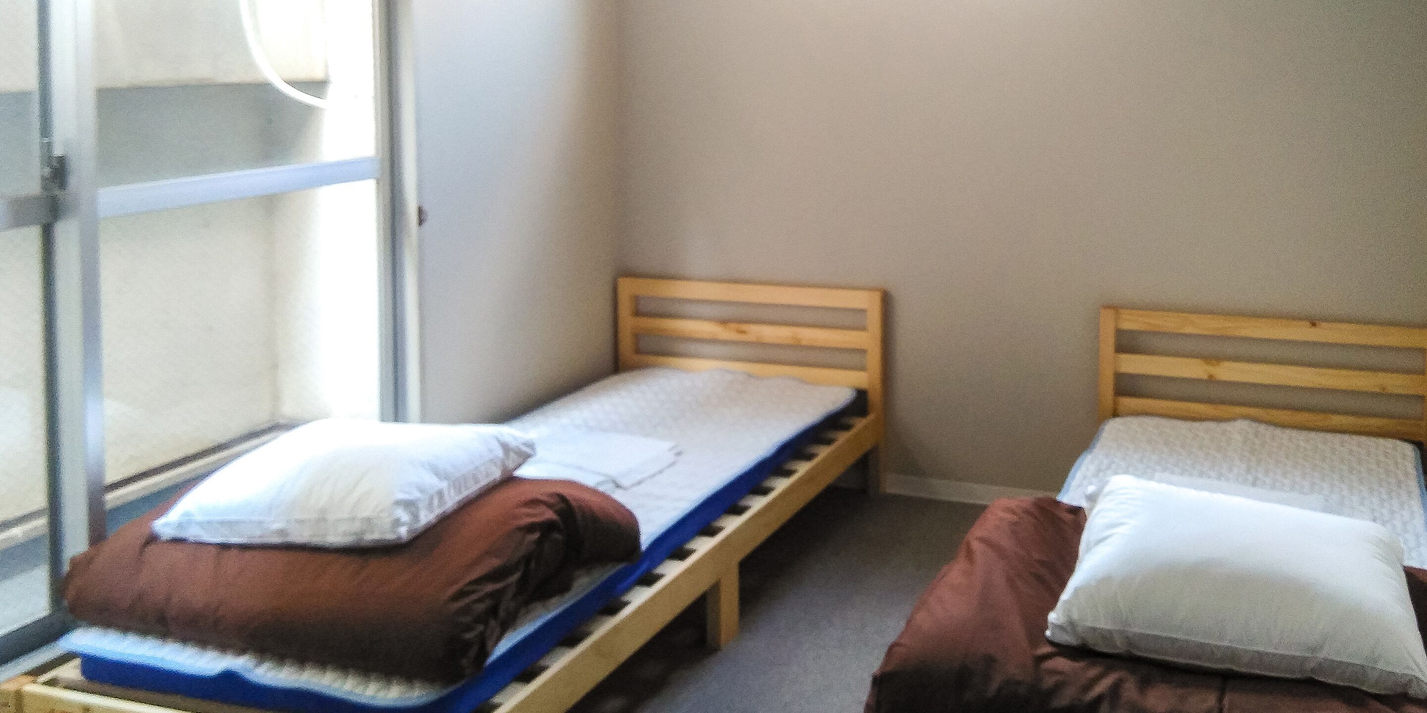 Two single beds with wooden frames and mattresses, a window with blinds, and a plain wall.