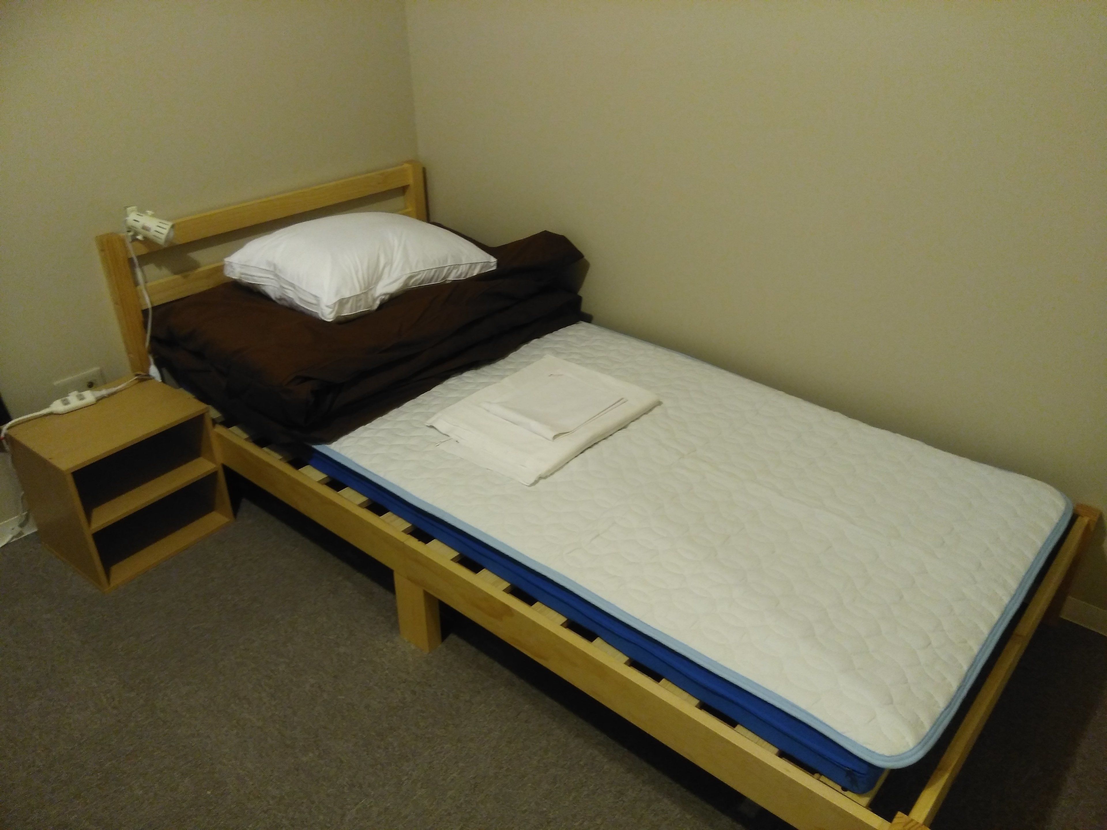 Una cama individual con estructura de madera, colchón blanco y manta marrón. Junto a la cama, una mesita de noche de madera con un enchufe y un tapón.