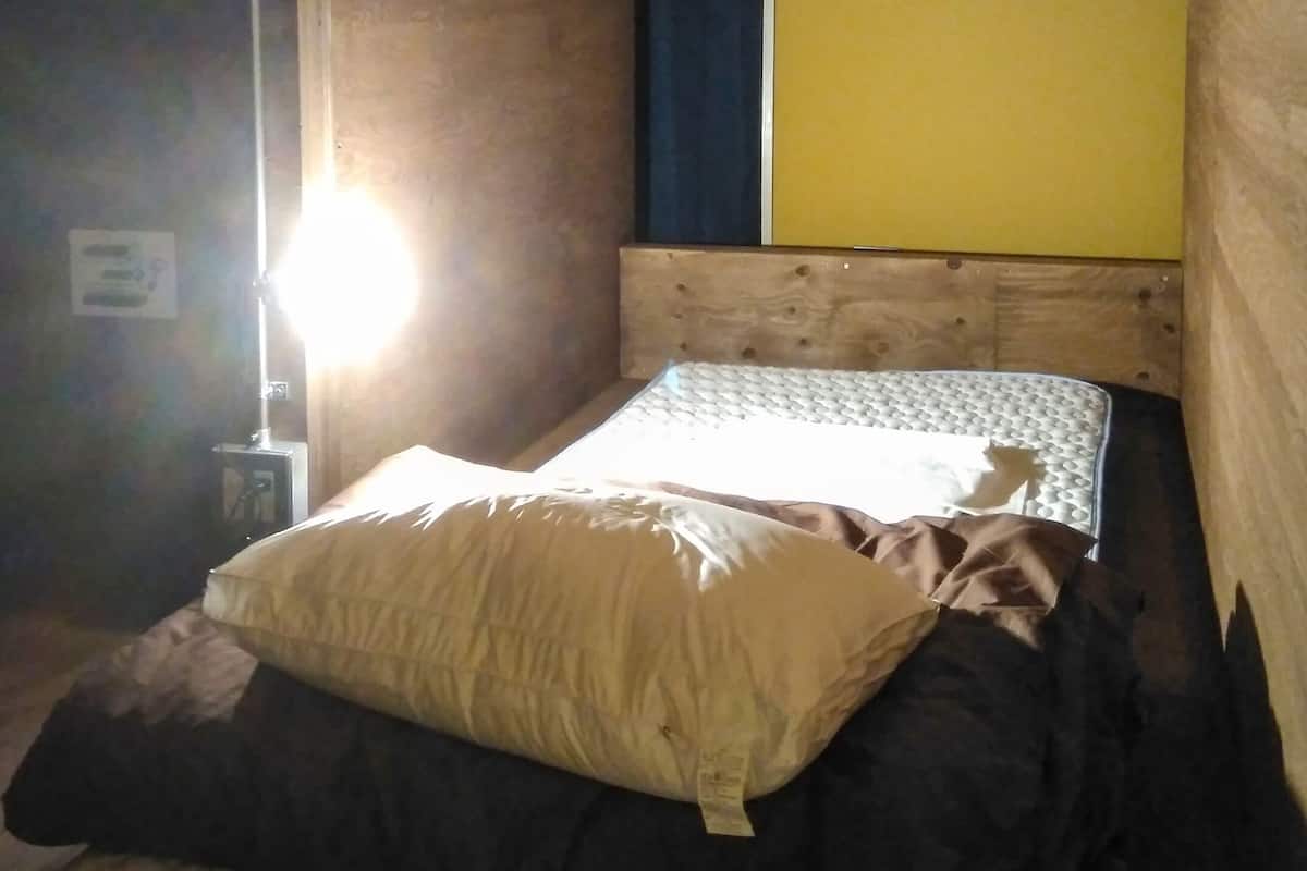 Una habitación pequeña y tenue, con una cama individual, una mesita de noche y un panel azul y amarillo en la pared.
