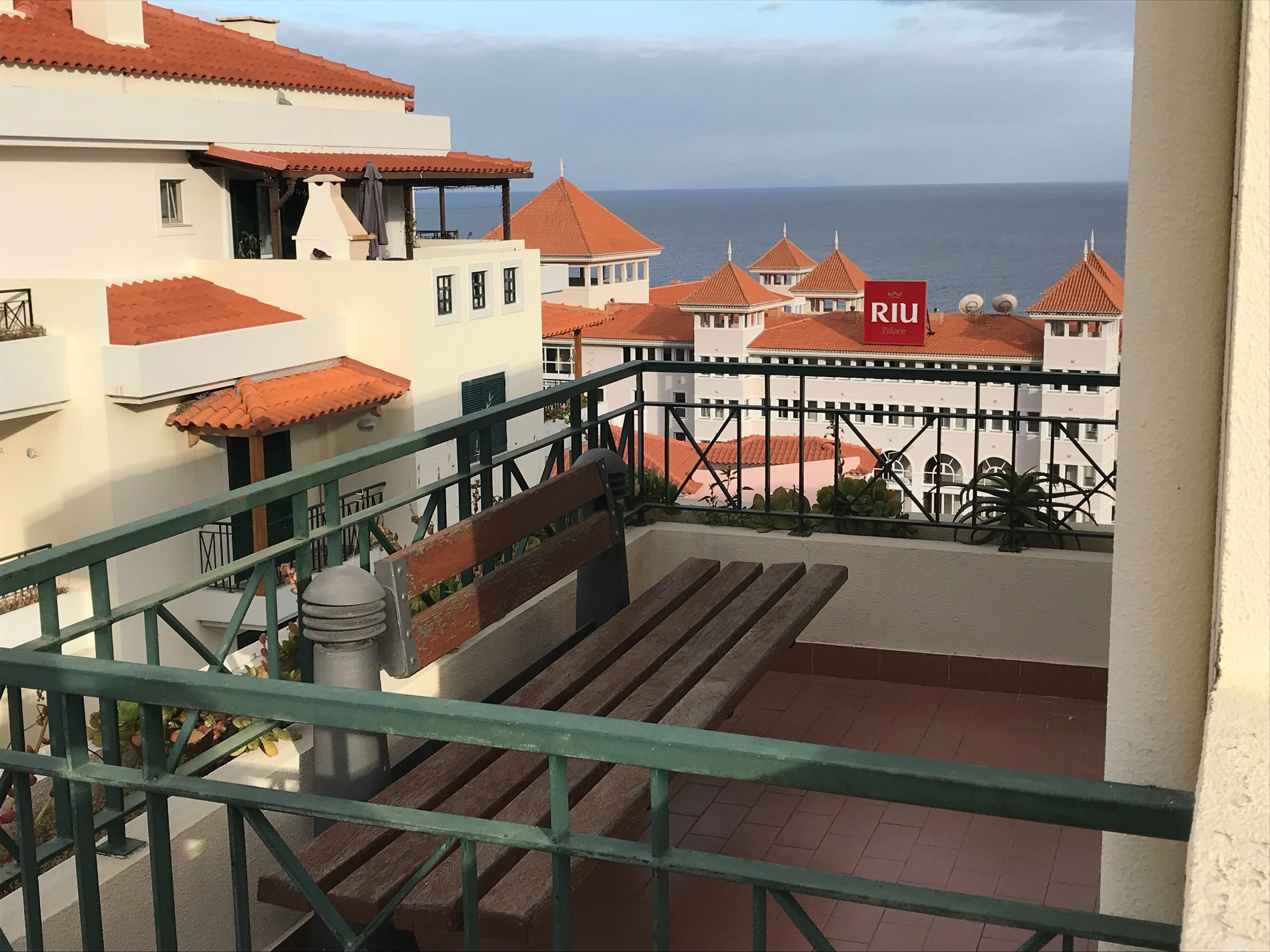 Un balcon donnant sur une zone côtière, avec des bâtiments aux toits recouverts de tuiles rouges et une enseigne indiquant « RIU Palace ».