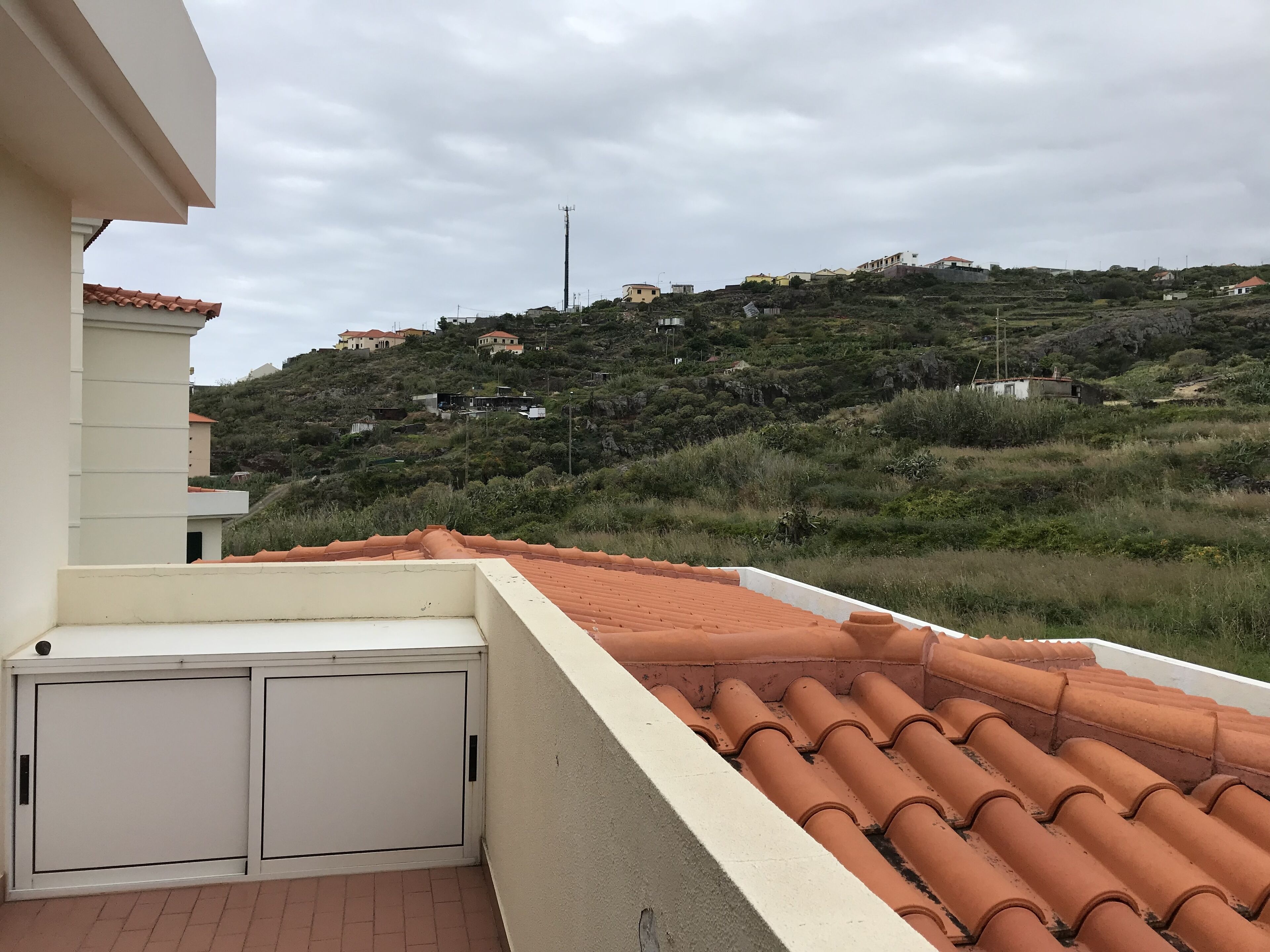 A rooftop terrace with terracotta tiles, a white wall, and a view of a hillside with buildings.