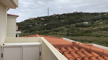 A rooftop terrace with terracotta tiles, a white wall, and a view of a hillside with buildings.