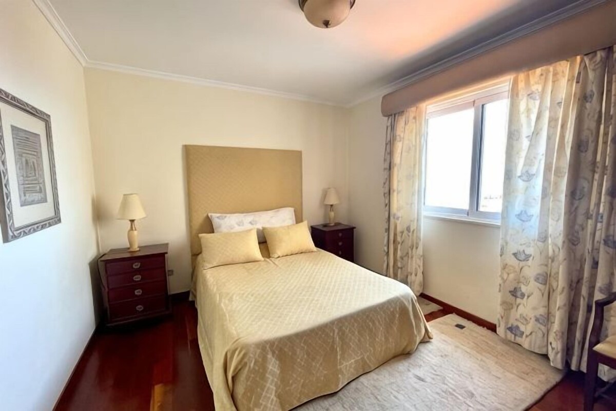 Une chambre à coucher avec un lit, deux tables de chevet, une fenêtre avec des rideaux et un tableau encadré au mur.