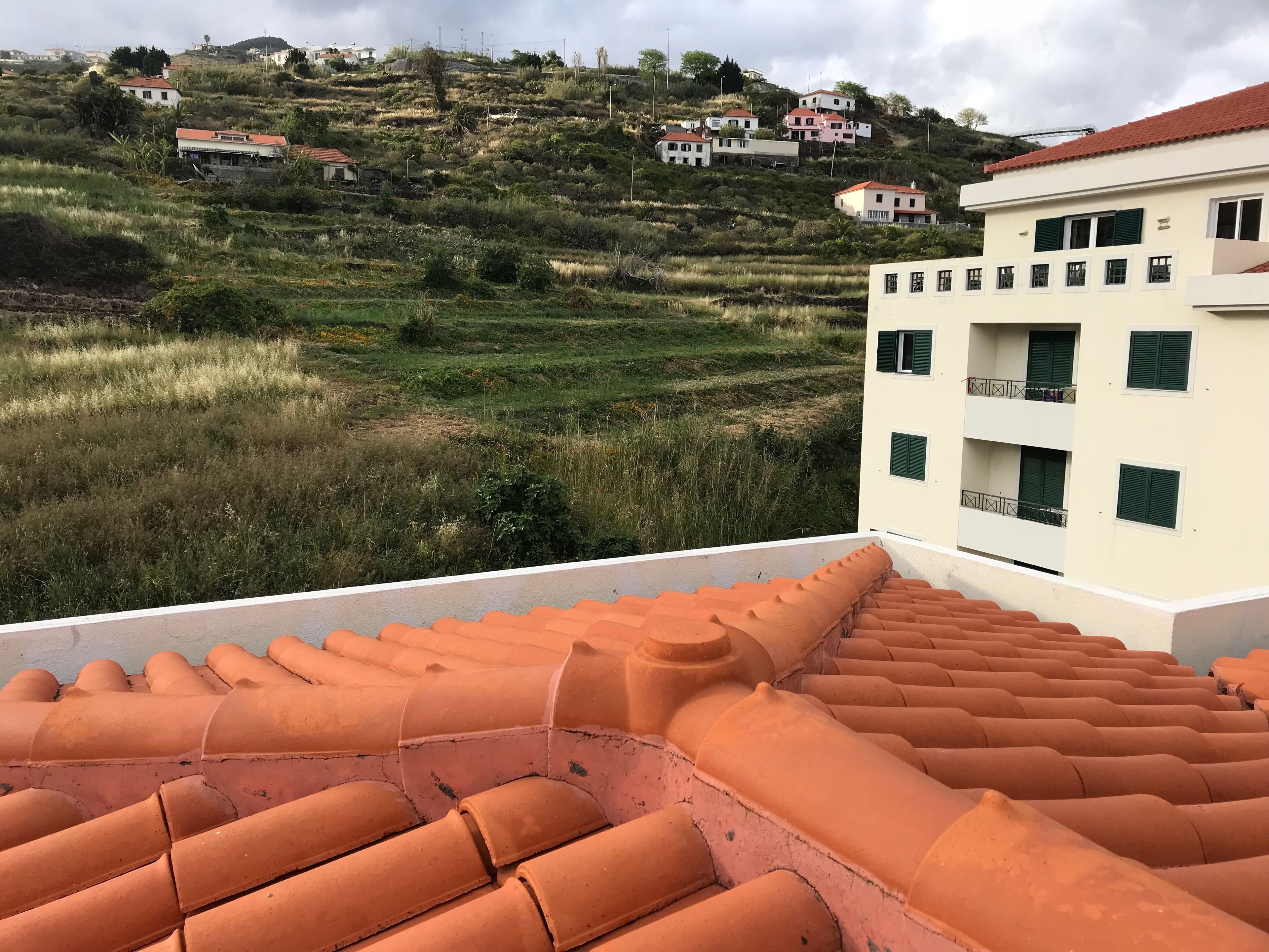 A rooftop terrace with terracotta tiles, overlooking a hilly area with houses and greenery.